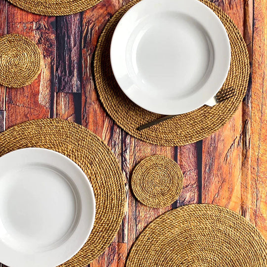 A beautifully set dining table featuring round handwoven placemats and coasters in natural tones, paired with white ceramic plates on a rustic wooden surface.